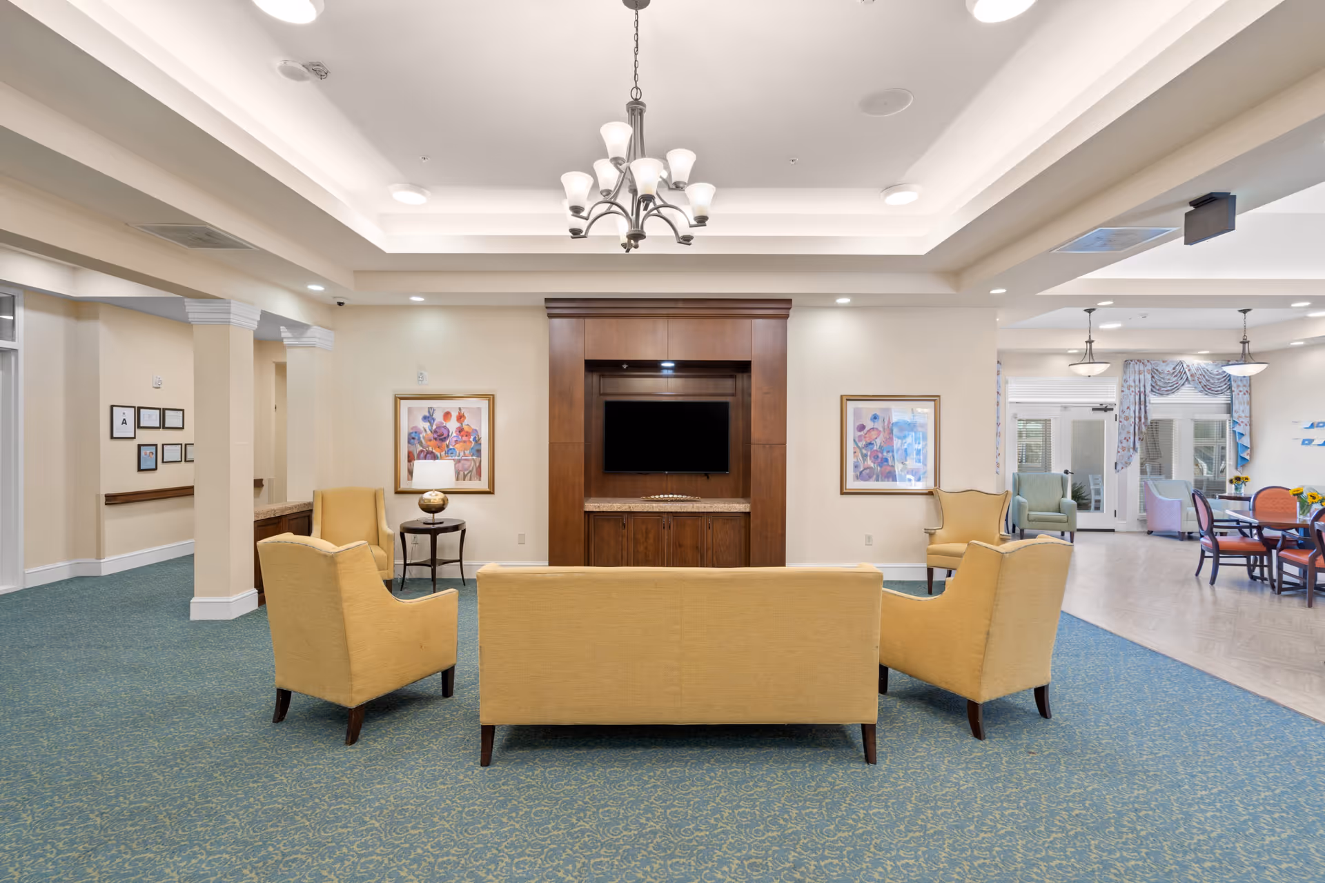A senior living facility common area with a seating arrangement consisting of a yellow sofa and two matching armchairs facing a wall-mounted flat screen TV set in a wooden entertainment center. The room has blue patterned carpet, beige walls, two framed floral paintings, and a chandelier hanging from a tray ceiling. In the background, there is a dining area with tables, chairs, and large windows with floral curtains.