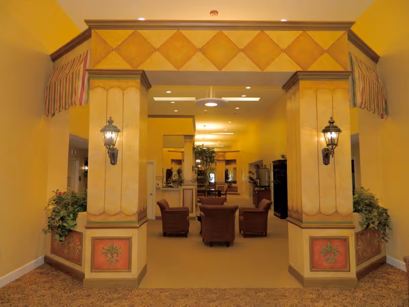 Interior view of a senior living facility lounge area with yellow walls, decorative columns, wall-mounted lantern-style lights, and several cushioned armchairs arranged around a table. There are plants on either side of the columns and ceiling lights illuminating the space.