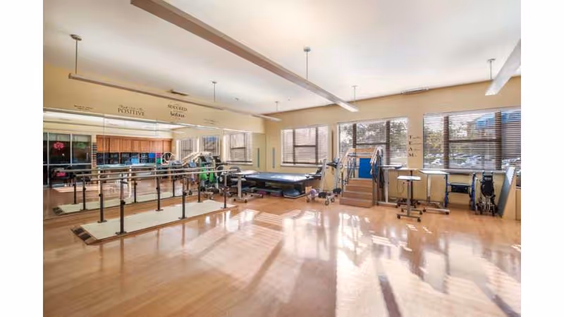A spacious physical therapy room with wooden floors and large windows letting in natural light. The room contains parallel bars for walking exercises, a therapy table, stairs for rehabilitation, various exercise equipment, and wheelchairs. The walls have motivational words like 'Positive' and 'Succeed'.