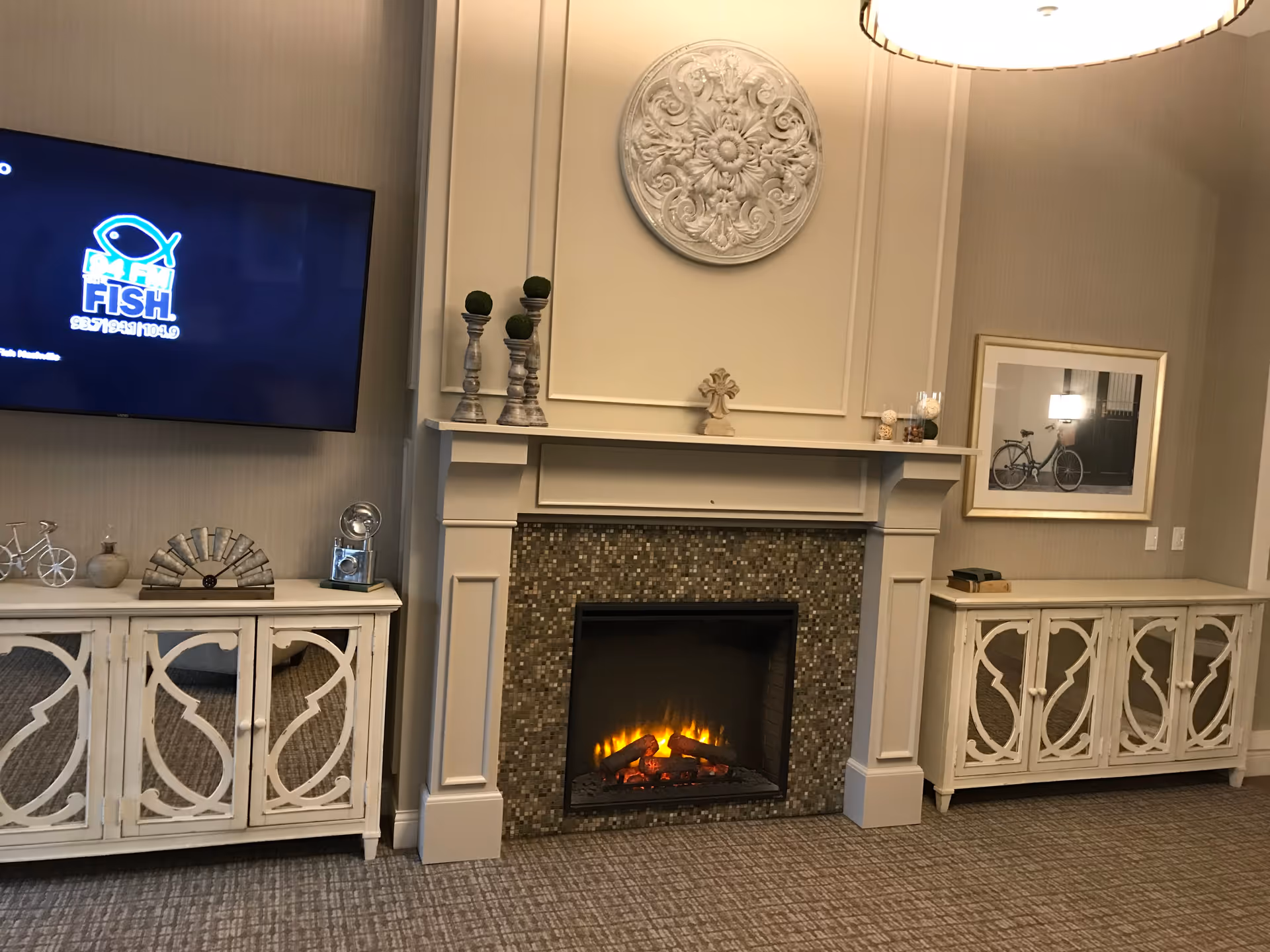 A cozy living room area featuring a lit electric fireplace with a decorative white mantel and mosaic tile surround. On either side of the fireplace are white wooden cabinets with intricate cut-out designs. Above the fireplace hangs a round ornamental wall decor, and to the right, a framed picture of a bicycle is mounted on the wall. A flat-screen TV is mounted on the left wall above one of the cabinets. The room has a warm, neutral color palette with carpeted flooring.