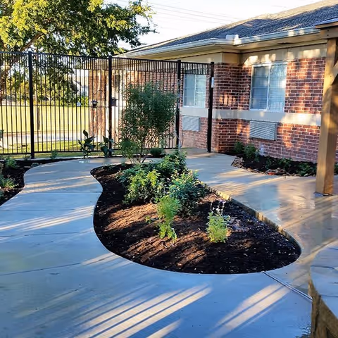 Curved concrete walkway surrounding a garden bed with small plants and shrubs, leading to a black metal gate attached to a brick building with windows.