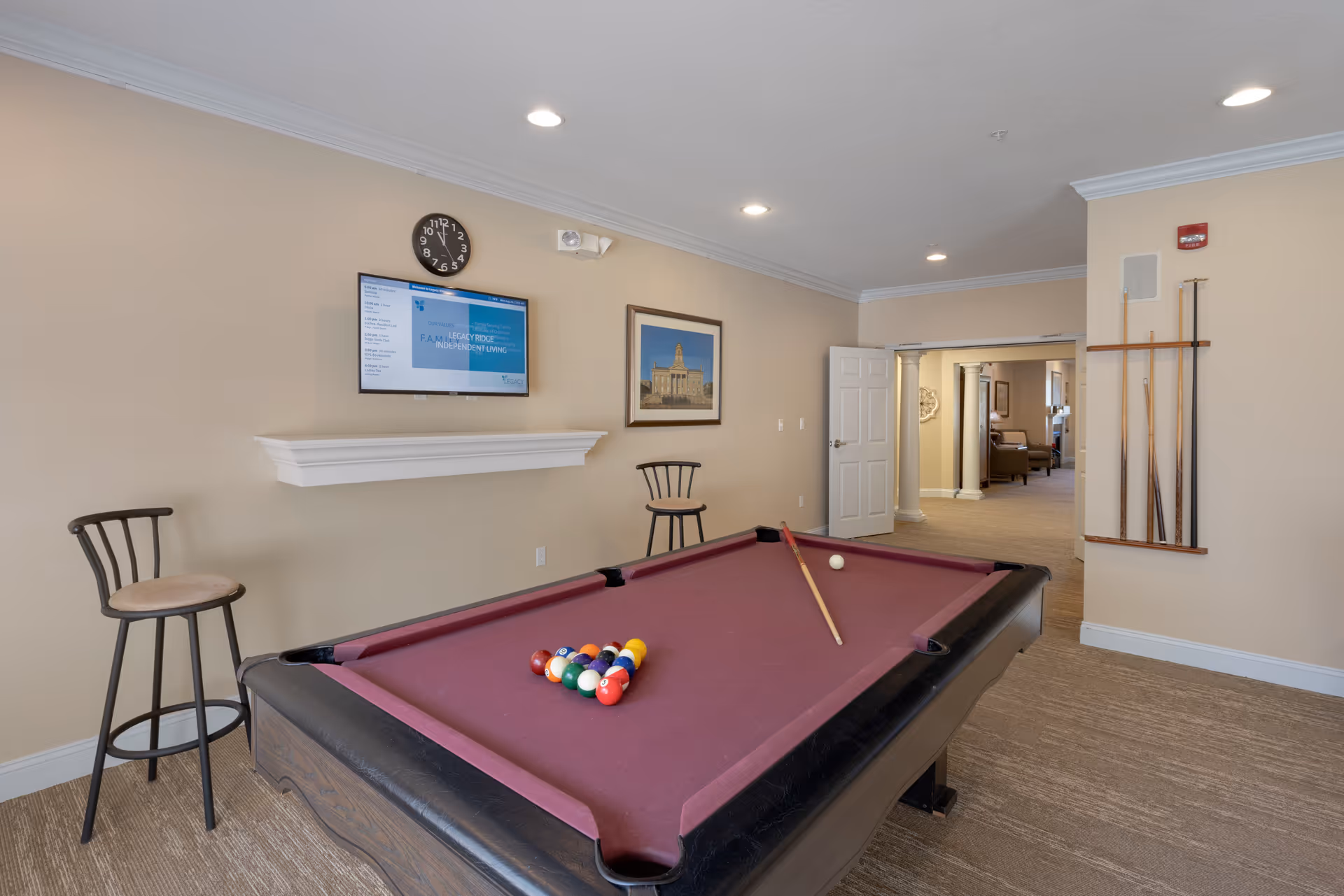 A recreation room with a maroon-felt pool table, cue rack, wall-mounted TV and two bar stools.