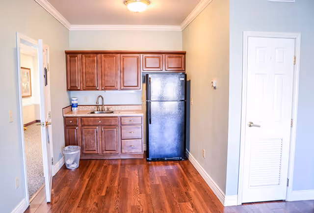 Small kitchenette with wooden cabinets, a sink, a black refrigerator, and hardwood floors in an apartment-style interior.