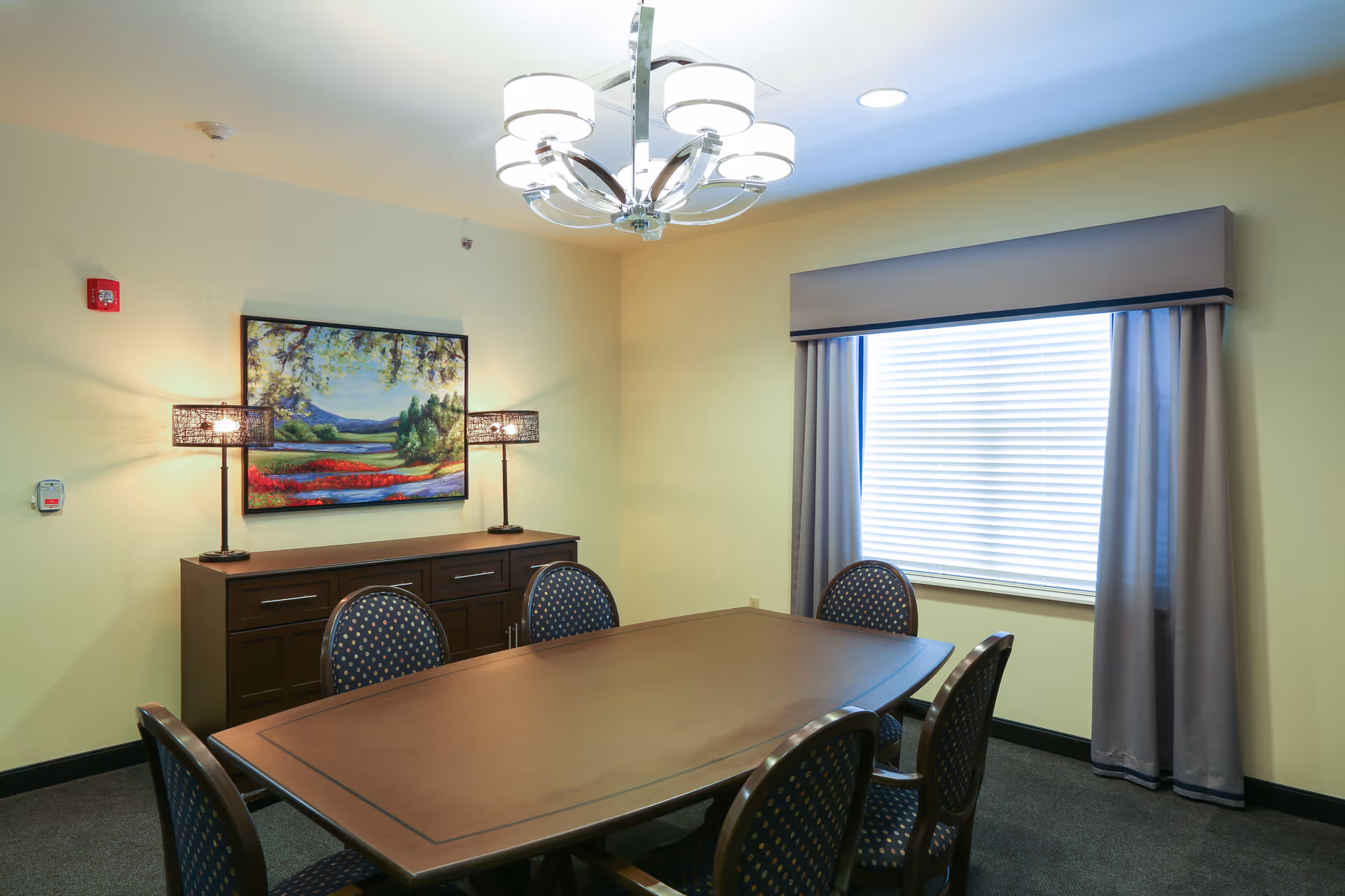 Dining room with a rectangular wooden table surrounded by chairs, a sideboard topped by a landscape painting and lamps, and a window with blinds and drapes.