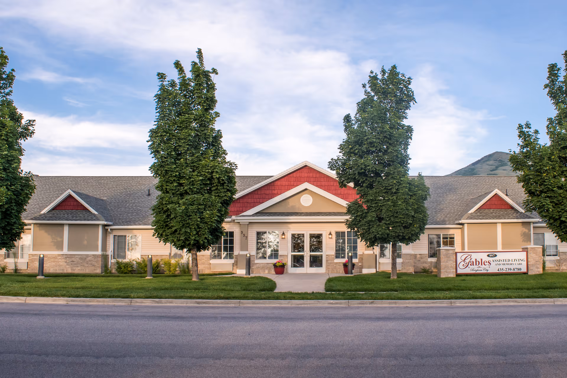 Front exterior view of The Gables Assisted Living of Brigham City building with a well-maintained lawn, several trees, and a clear sky in the background.