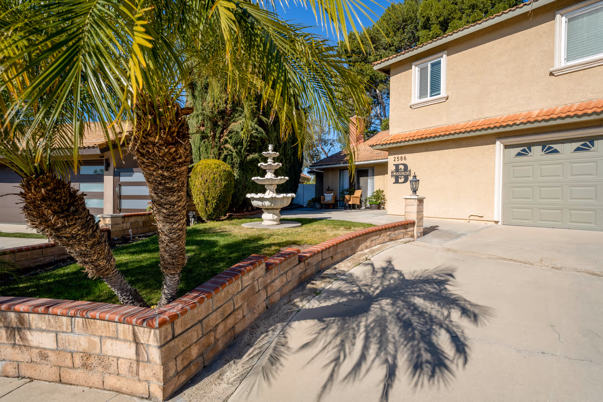 Exterior view of a two-story beige stucco building with a tiled roof, a green garage door, and a driveway. In front of the building is a small garden area with palm trees, a trimmed bush, and a white decorative fountain. The building has a sign that reads 'Blessings' with the number 2586 above it. The sky is clear and blue.