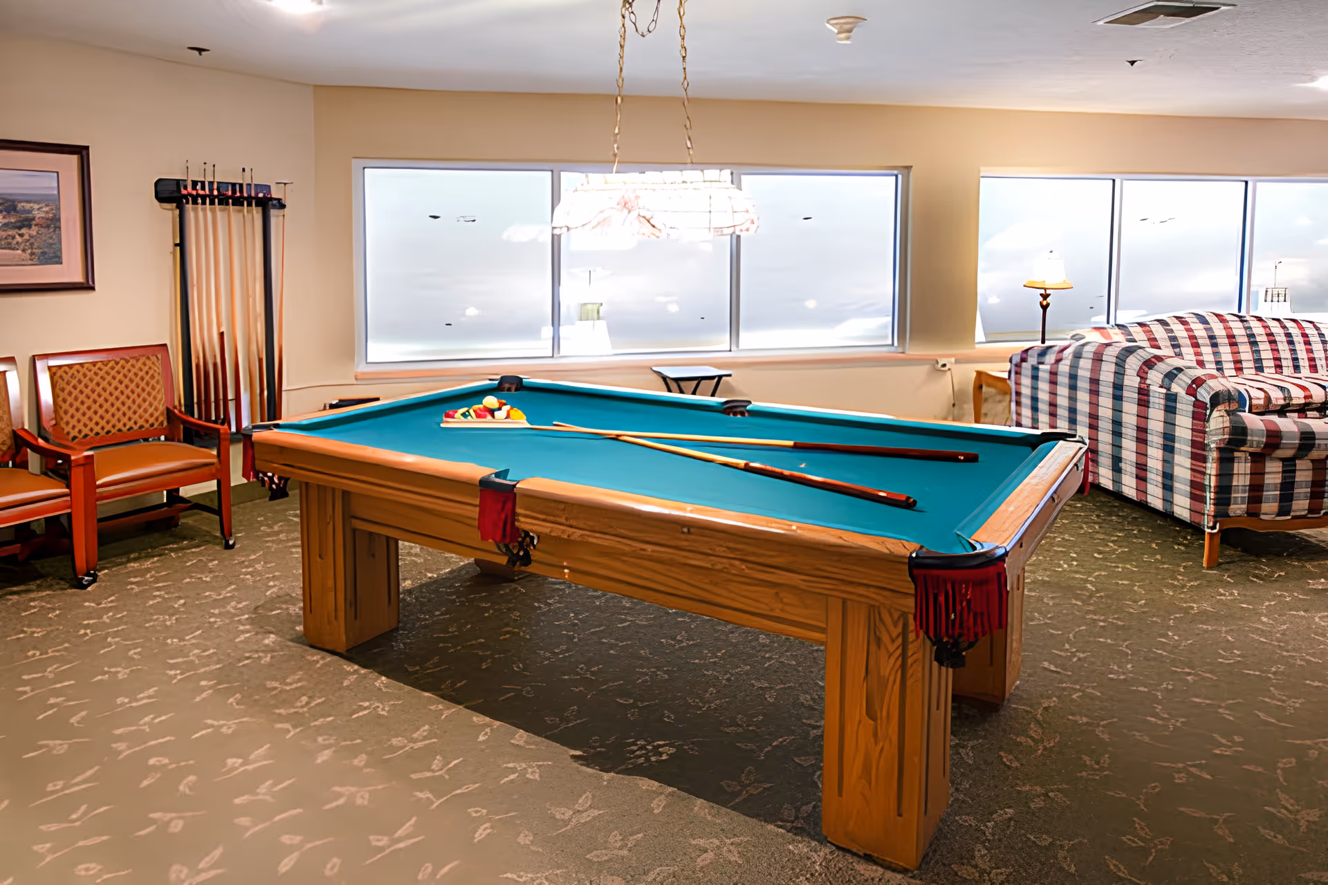 A recreational room with a pool table in the center, pool cues resting on the table, and a rack of additional cues against the wall. There are two wooden chairs with patterned cushions to the left and a plaid upholstered couch to the right near large windows letting in natural light. A hanging light fixture is above the pool table.
