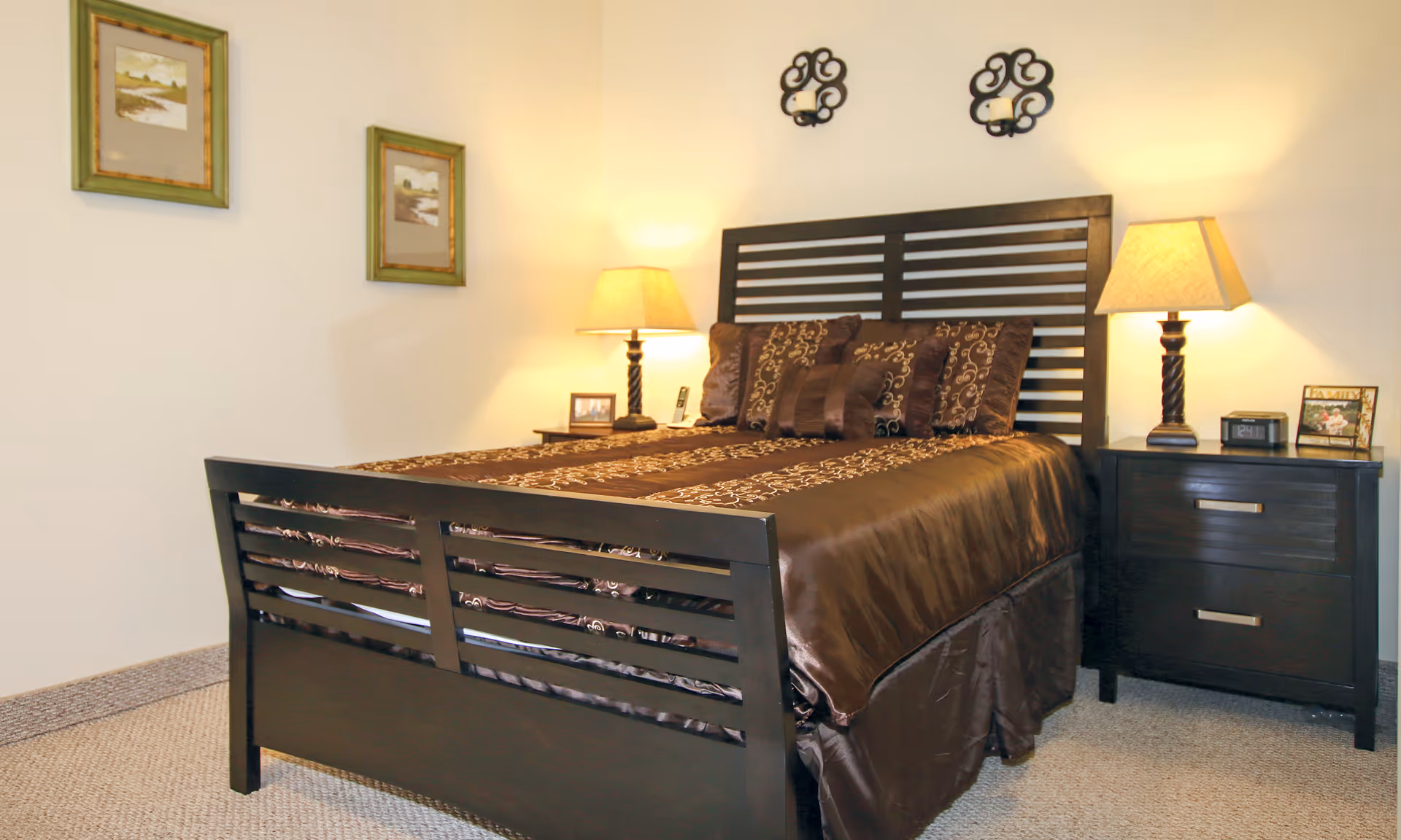 Bedroom with a dark wooden bed dressed in brown bedding, two nightstands with lamps, and wall decor.