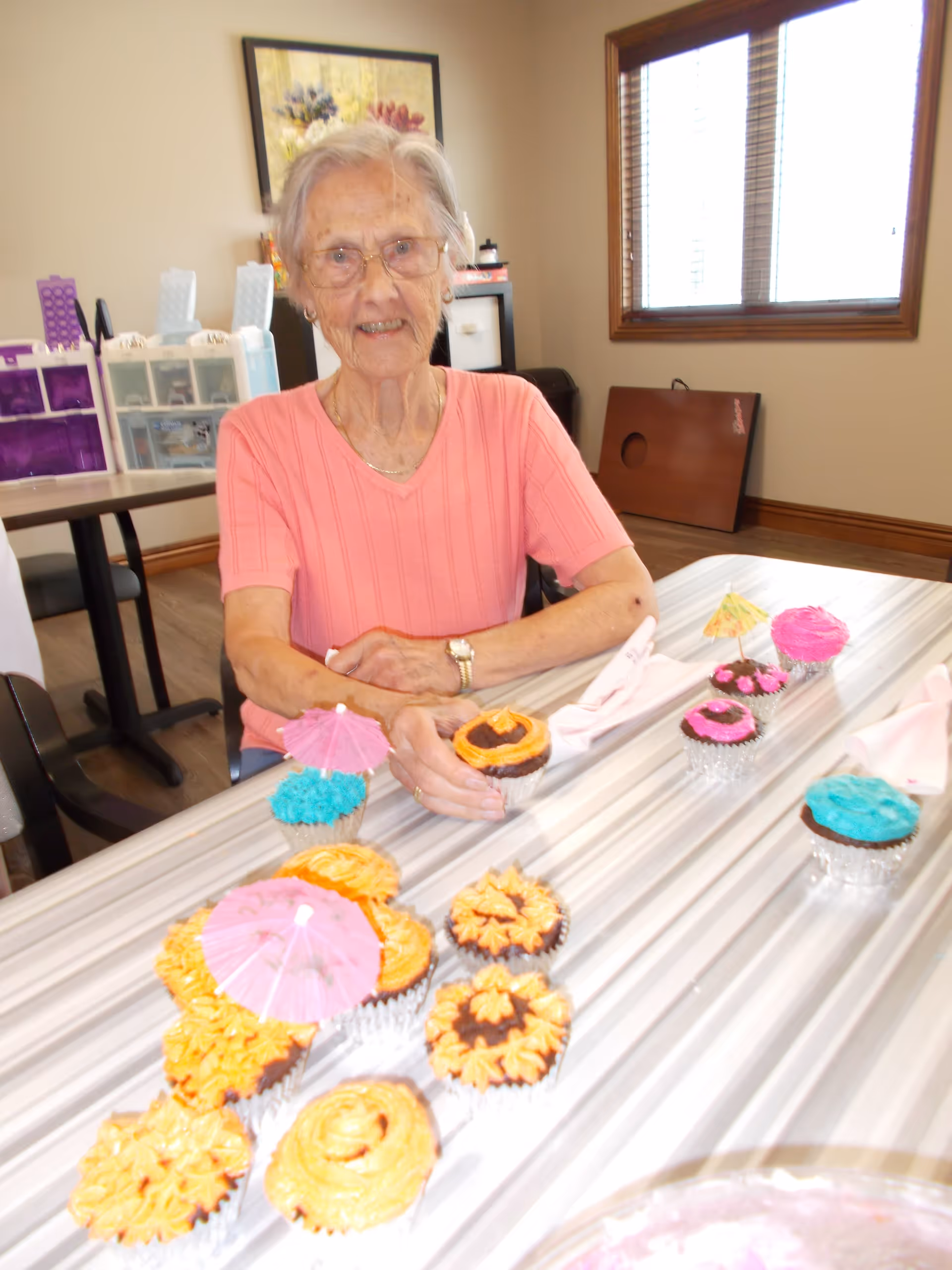 An elderly woman wearing glasses and a pink short-sleeve shirt sits at a table with a variety of colorful decorated cupcakes, some with small paper umbrellas. The room has a window with blinds, a framed floral picture on the wall, and storage containers in the background.