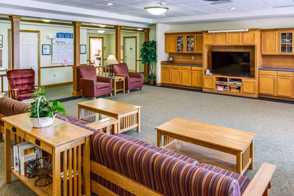 A spacious living room area in a senior living facility featuring a striped couch, wooden coffee tables, two maroon armchairs, a wooden entertainment center with a large flat-screen TV, and a potted plant. The room has beige walls, carpeted flooring, and a ceiling with recessed lighting. A bulletin board labeled 'ACTIVITIES' is visible on the wall in the background.