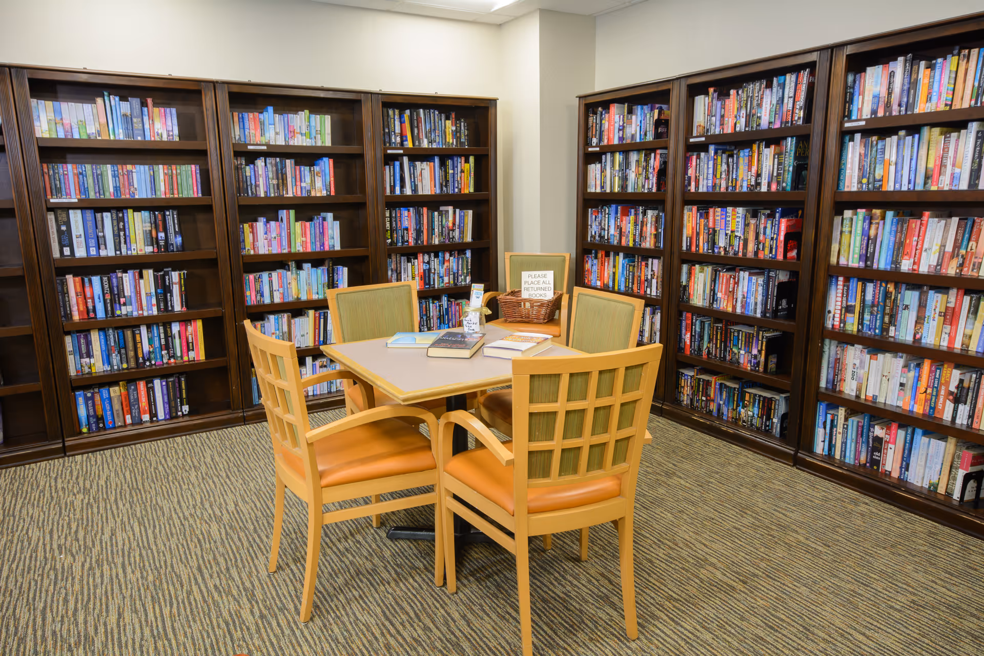 A cozy library room with tall bookshelves lining the walls and a square table with four chairs in the center.
