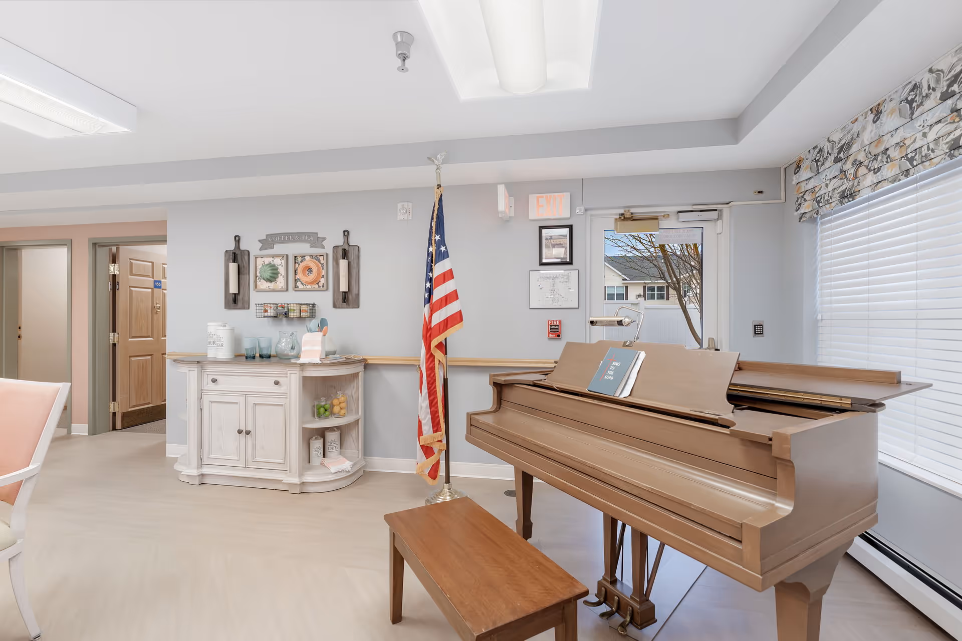 Interior room with a wooden piano and bench near a window with blinds. An American flag stands next to a white cabinet with decorative items on and above it. The room has light-colored walls and flooring, with a door and hallway visible in the background.