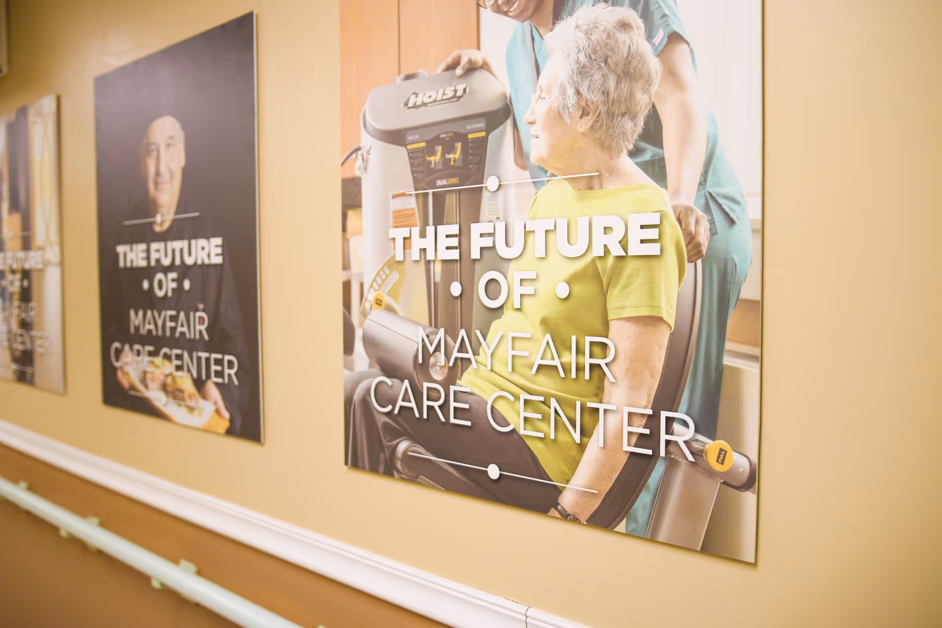 A hallway wall with large posters featuring elderly individuals and caregivers at Mayfair Care Center. One poster shows an elderly woman using exercise equipment with a caregiver assisting her, and another poster shows an elderly man holding a plate of food. Both posters have the text 'THE FUTURE OF MAYFAIR CARE CENTER'.