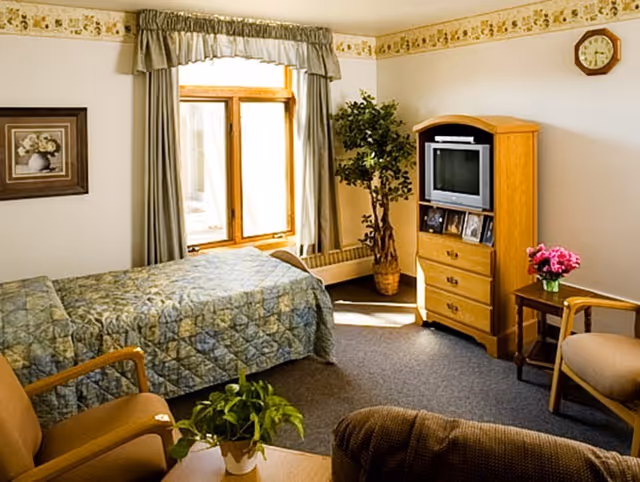 A cozy bedroom in a senior living facility featuring a single bed with a patterned quilt, a wooden TV cabinet with a television and framed photos, a small side table with a vase of pink flowers, two armchairs, a potted plant on a coffee table, a window with curtains, and a wall clock.