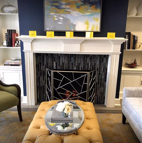 Cozy living room scene with a white mantel fireplace and dark tile surround, a tufted yellow ottoman topped with a mirrored tray and flowers, artwork above and built-in shelves on either side.