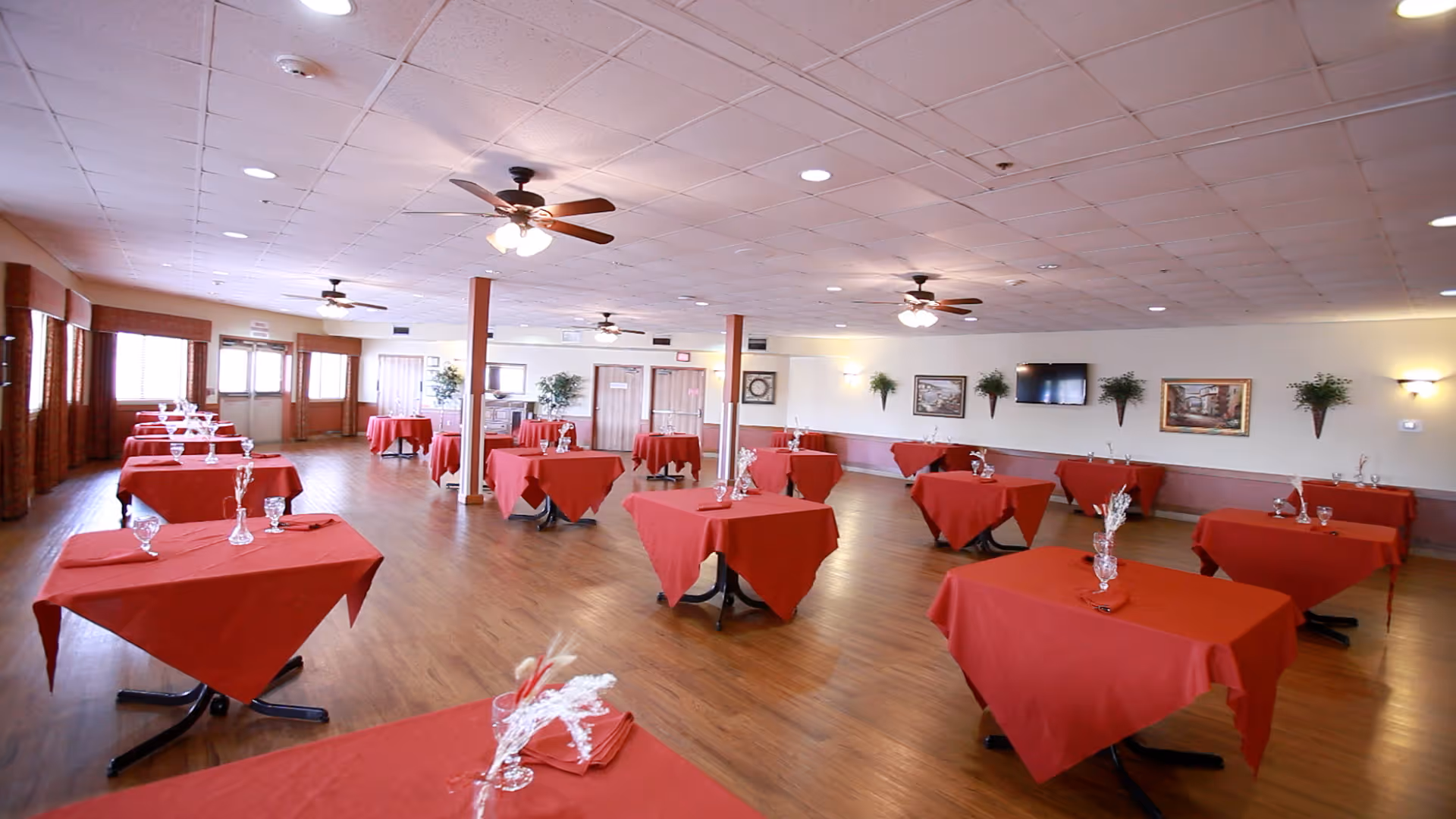 A spacious dining room with multiple square tables covered in red tablecloths, each set with glassware and napkins. The room has wooden floors, ceiling fans with lights, wall-mounted plants, framed pictures, and a flat-screen TV on the wall. Large windows with brown curtains allow natural light to enter.