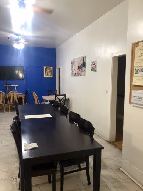 Interior view of a dining area in a senior living facility with a long black table and several black chairs. In the background, there is a smaller wooden dining table with chairs against a blue accent wall with a mounted flat-screen TV. The walls are decorated with framed pictures and a bulletin board is visible near a doorway.