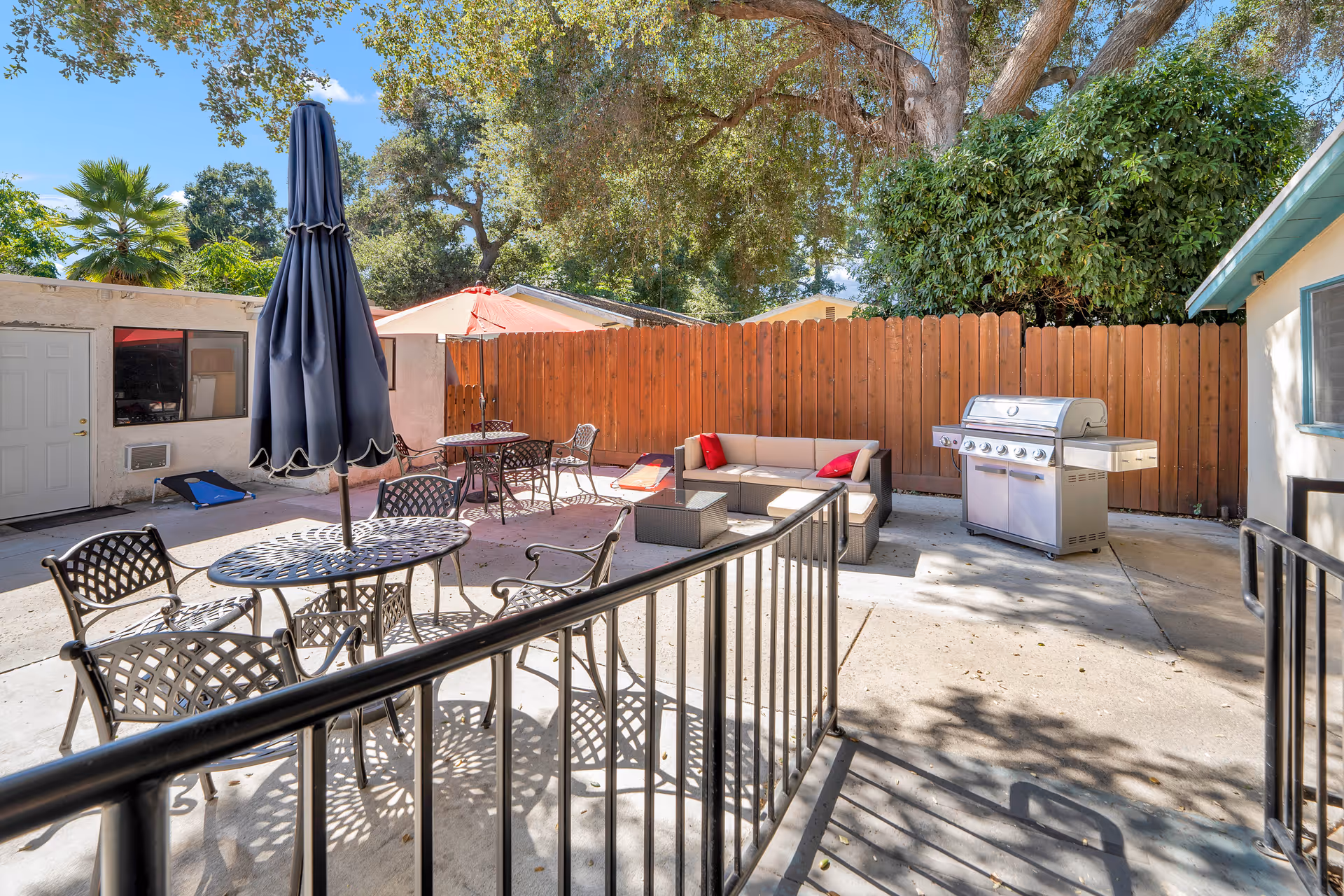 Outdoor patio with metal tables and chairs, a closed umbrella, lounge seating and a stainless steel grill beside a wooden fence under trees.