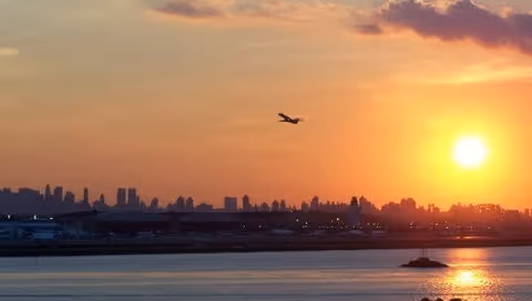 A sunset view over a body of water with a city skyline in the distance and an airplane flying in the sky.