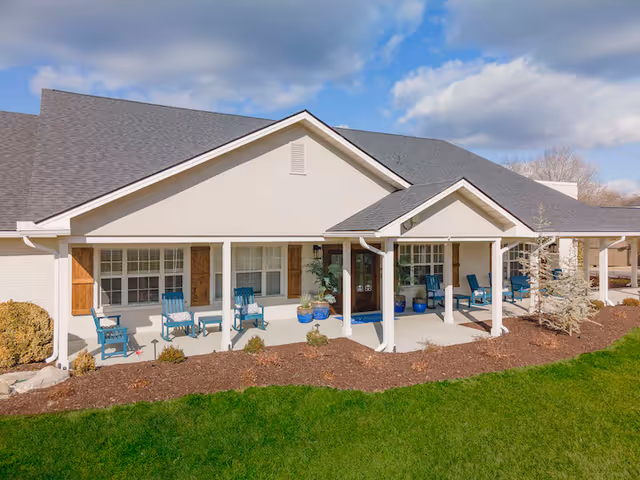 Exterior view of a single-story building with a covered porch featuring several blue chairs and potted plants. The building has a light-colored facade with wooden shutters on the windows and a dark shingled roof. The foreground shows a well-maintained lawn and landscaped garden beds under a partly cloudy sky.