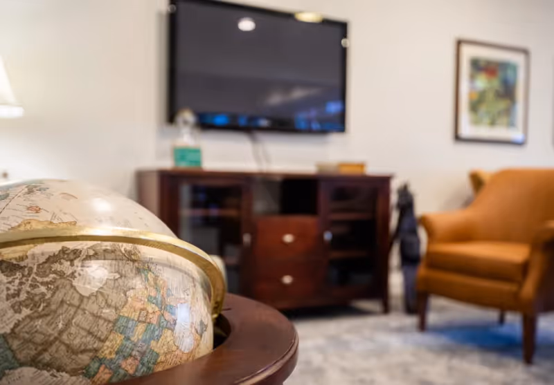 Globe in the foreground with a wall-mounted TV, wooden media cabinet, and an upholstered armchair in a living room.