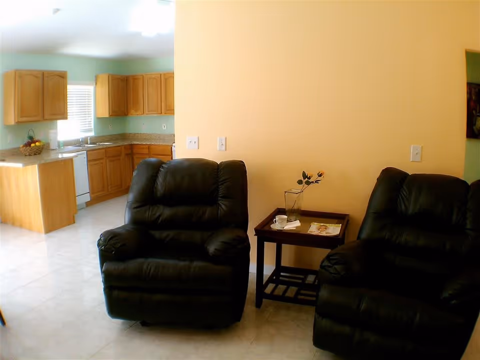 Two black leather recliners and a side table in a living area with a view into a kitchen with wooden cabinets.