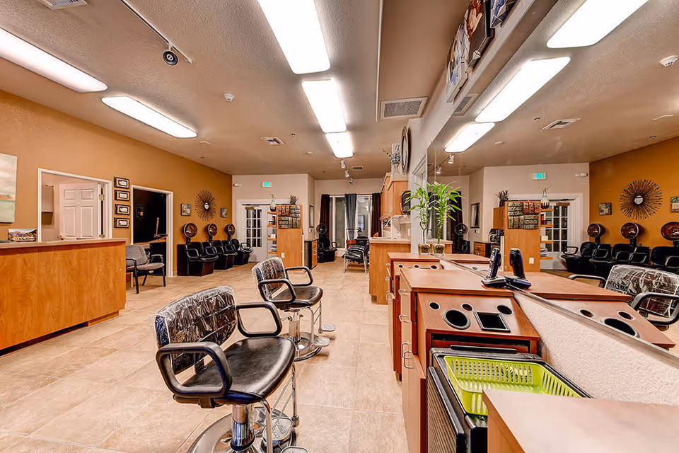Interior view of a salon area with multiple black salon chairs, mirrors, and wooden cabinetry. The room has beige walls, tiled floors, and bright overhead lighting. There are additional black chairs lined up against the far wall and decorative wall art.