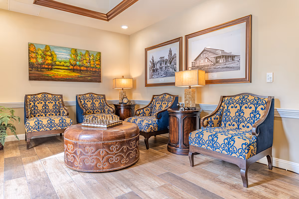 A cozy seating area in a senior living facility featuring four upholstered armchairs with blue and beige patterned fabric arranged around a round brown leather ottoman with decorative nailhead trim. Two wooden side tables with table lamps are placed between the chairs. The walls are adorned with framed artwork, including a colorful landscape painting and two black and white photographs of buildings. The room has wooden flooring and soft, warm lighting.