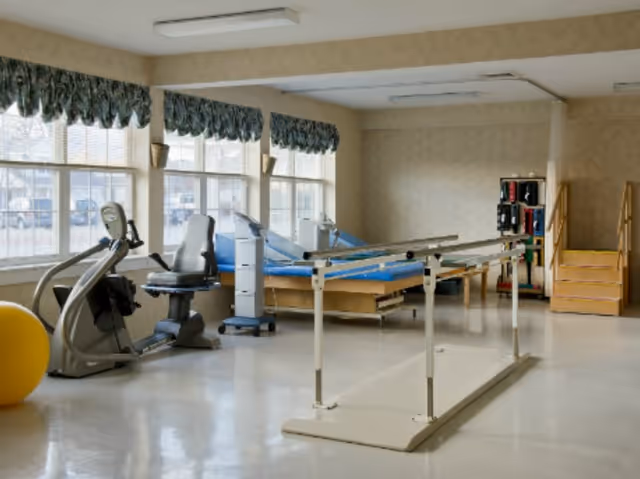 Physical therapy room with exercise equipment including a stationary bike, parallel bars for walking practice, therapy beds, a yellow exercise ball, and a set of stairs with handrails. Large windows with valances allow natural light into the room.
