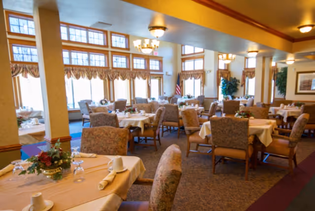 A bright dining room with several set tables, upholstered chairs, large windows, and ceiling chandeliers.