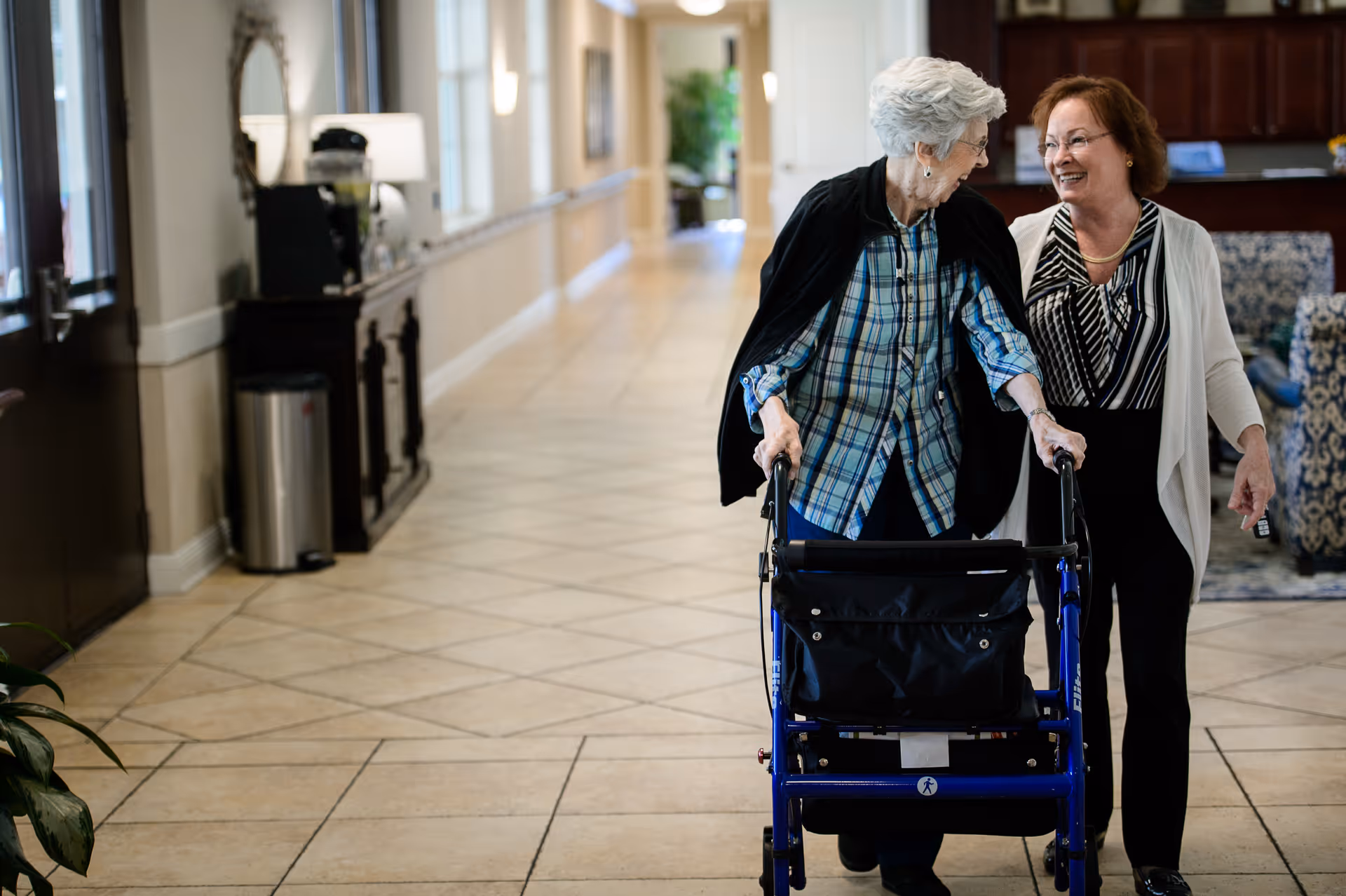 An elderly woman using a walker is accompanied by a smiling caregiver as they walk together down a tiled hallway in a senior living facility.