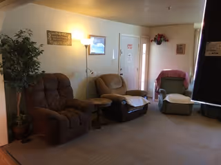 Three recliner chairs and small tables arranged in a carpeted living room area with a floor lamp, potted plant, and an entrance door.
