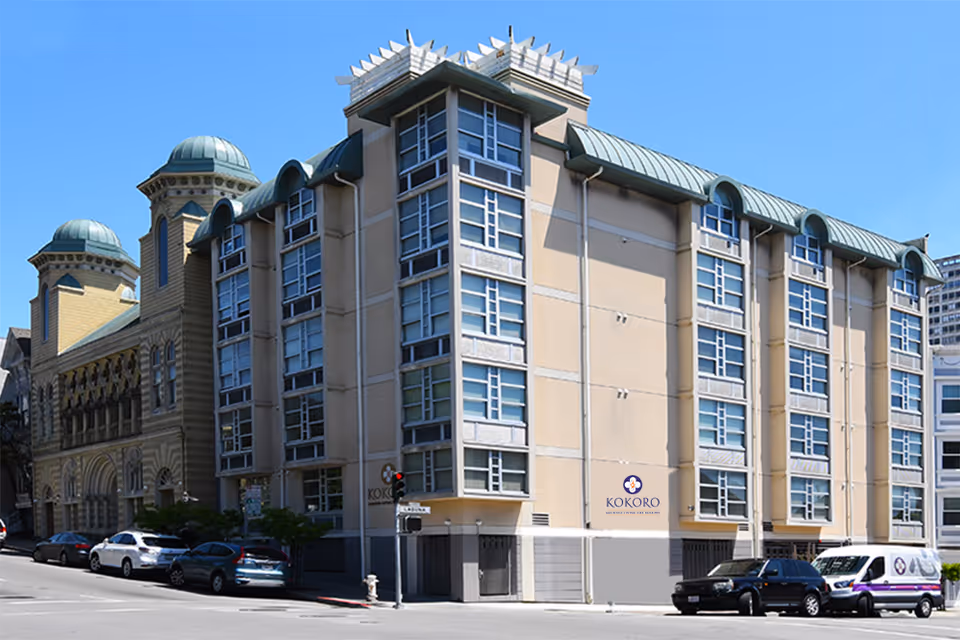 Exterior view of a multi-story beige building with many windows under a clear blue sky. The building has a sign that reads 'Kokoro Assisted Living Inc' and is located on a street corner with several parked cars along the street.