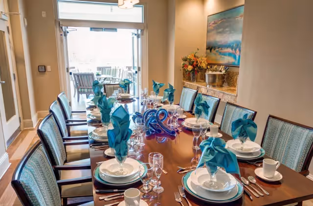 A formal dining room with a long wooden table set for a meal, featuring white plates, cups, silverware, wine glasses, and blue folded napkins. The room has upholstered chairs around the table, a sideboard with a floral arrangement and ice bucket, a large painting on the wall, and glass doors leading to an outdoor patio with seating.