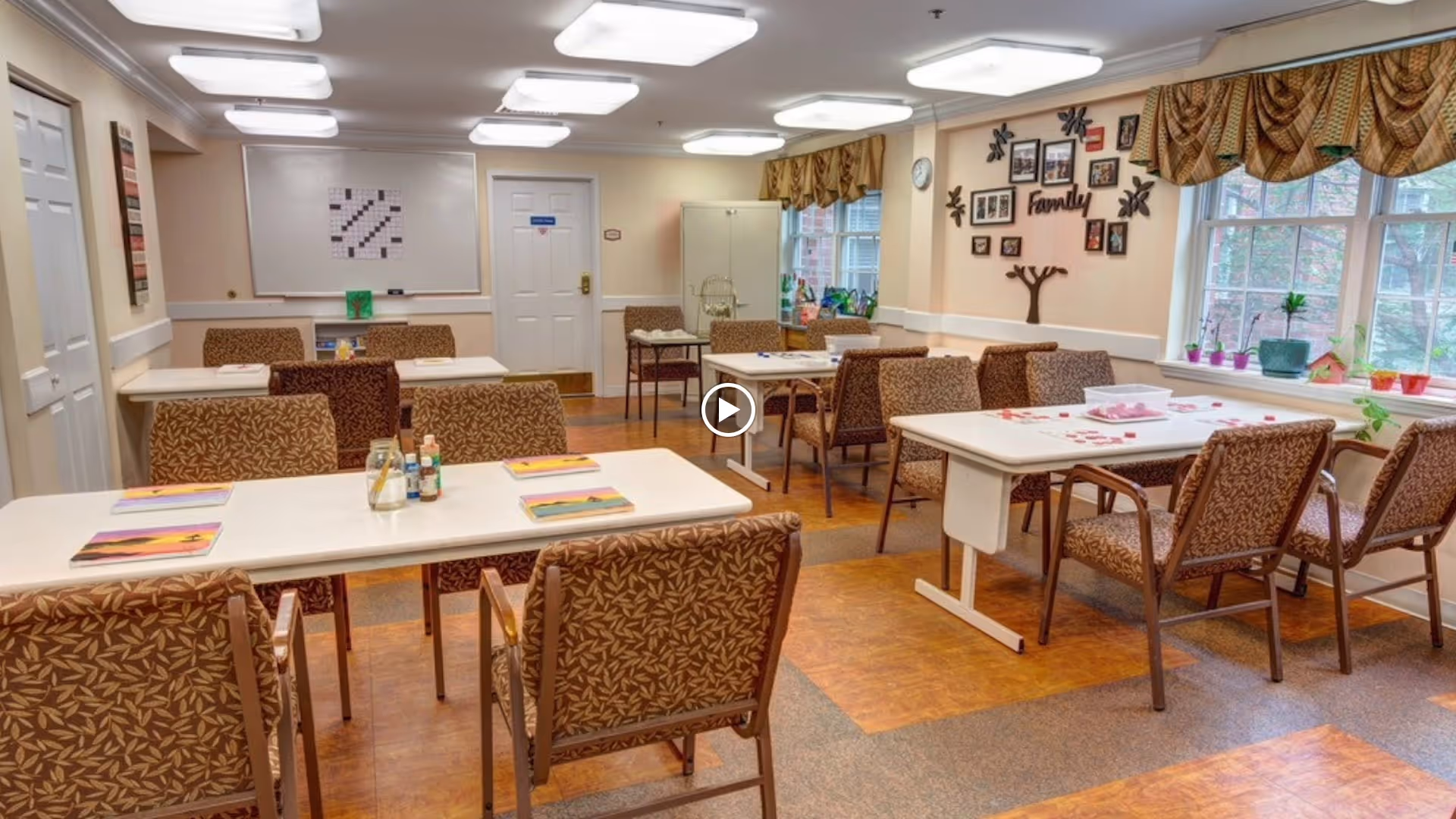 A well-lit room with multiple tables and chairs arranged for group activities or dining. The tables have books, bottles, and some game pieces on them. The walls are decorated with family-themed photo frames and a tree-shaped wall art. Large windows with patterned curtains allow natural light to enter the room.