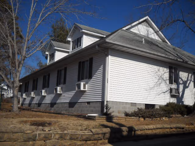 White two-story residential building with dormer windows, multiple window air conditioners, and a stone retaining wall under a clear blue sky.