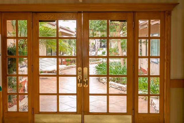 Wooden framed glass double doors leading to an outdoor garden area with tiled flooring, green plants, trees, and a stone-bordered walkway.