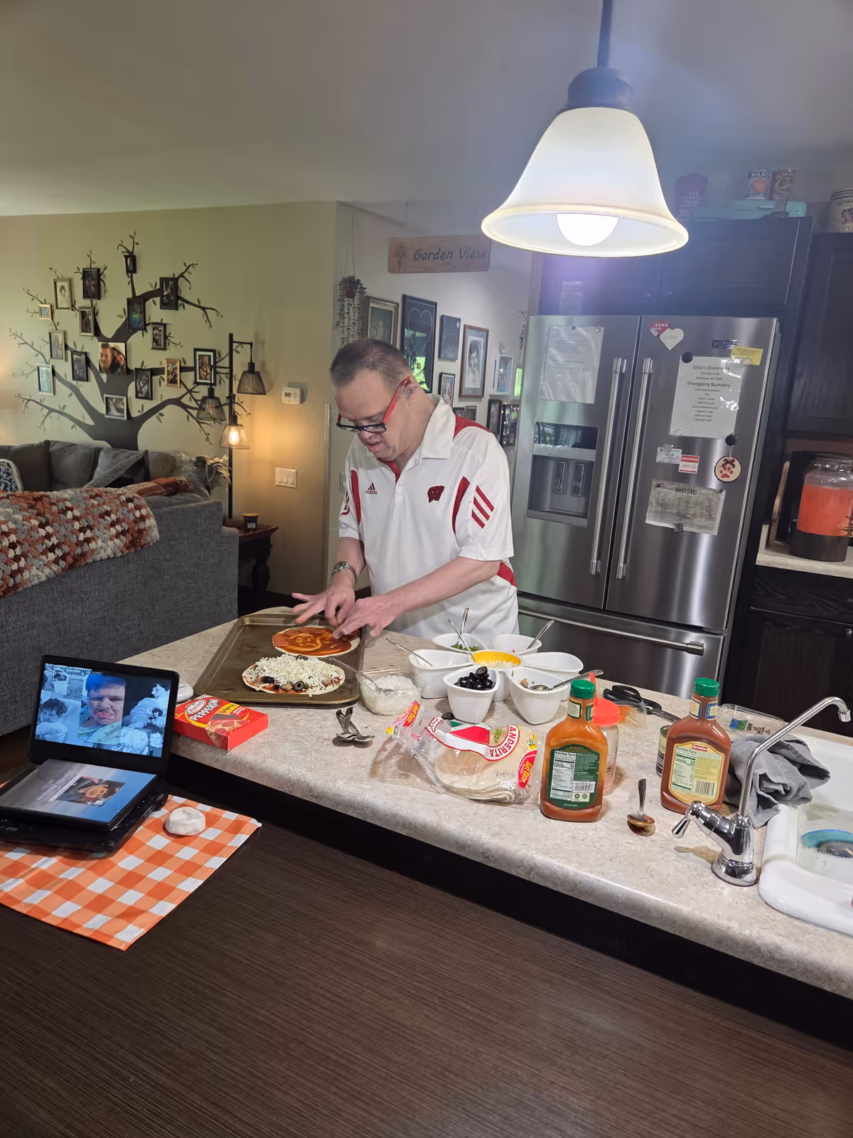 A man wearing glasses and a white and red shirt is preparing pizzas on a kitchen counter. Various pizza toppings and ingredients are spread out on the counter, including olives, cheese, and sauce. In the background, there is a stainless steel refrigerator with papers and magnets attached. The kitchen opens into a living room area with a gray couch and a wall decorated with a tree-shaped photo frame. A tablet on the counter displays a video call with multiple faces visible.