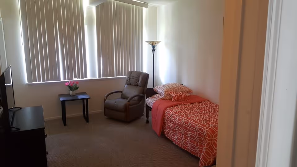 A small bedroom with a single bed covered in a red and white geometric patterned bedspread and pillows. Next to the bed is a tall floor lamp and a brown recliner chair. A small black side table with a vase of pink flowers is positioned near a window with vertical blinds. A TV is placed on a black dresser opposite the bed.