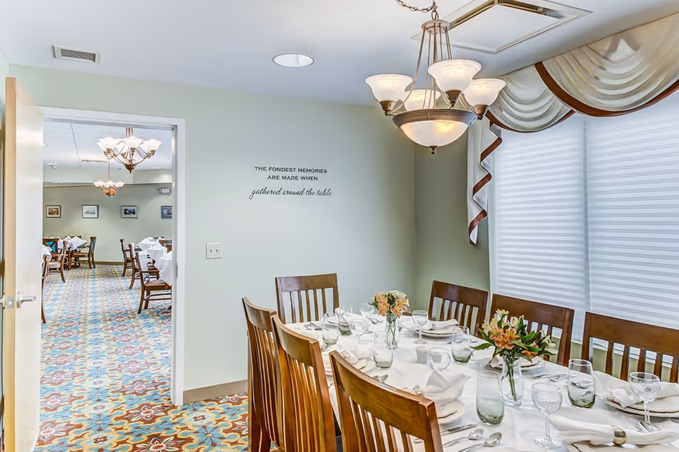 A dining area in a senior living facility with a wooden table set for a meal, including glasses, plates, napkins, and floral centerpieces. The room has a large window with white blinds and decorative valances. A chandelier hangs above the table. Through an open door, another dining room with multiple tables and chairs is visible. On the wall, there is a quote that reads, 'The fondest memories are made when gathered around the table.'
