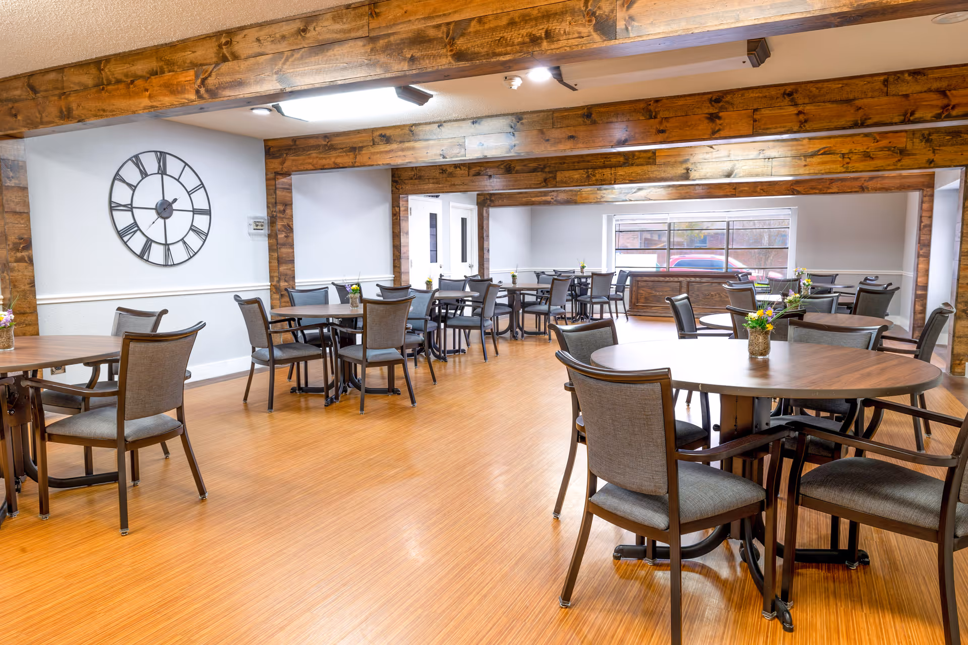 A spacious dining room with multiple round and rectangular wooden tables surrounded by cushioned chairs. The room features wooden beams on the ceiling and walls, a large clock on one wall, and a window letting in natural light. Small flower arrangements are placed on each table.