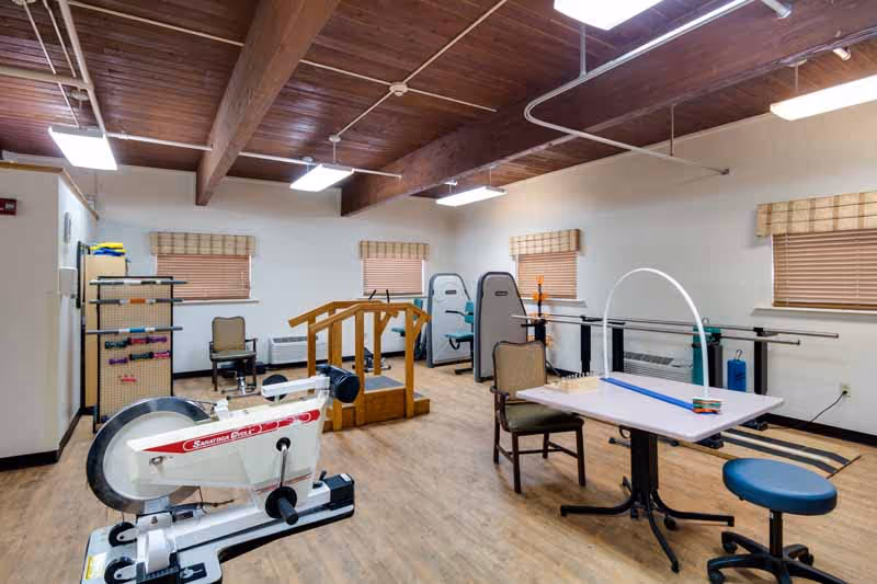 A rehabilitation or physical therapy room with exercise equipment including a stationary bike, parallel bars, a small wooden staircase for walking practice, and a table with chairs and therapy tools. The room has wooden beams on the ceiling, three windows with blinds, and bright overhead lighting.