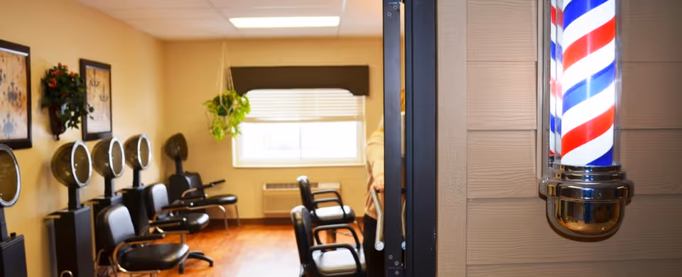 Interior view of a salon area in a senior living facility with several black salon chairs and hair dryers lined up against a beige wall. A window with blinds and a hanging plant is visible in the background. A traditional red, white, and blue barber pole is mounted on the exterior wall near the entrance.