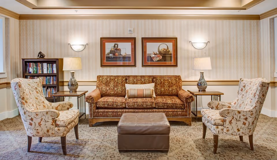 A cozy living room area with a patterned brown sofa, two floral upholstered armchairs facing each other, a brown leather ottoman in the center, two side tables each with a decorative lamp, a small bookshelf filled with books, and two framed pictures of baskets on the wall behind the sofa.