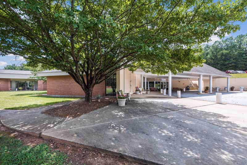 Exterior view of Seneca Health and Rehabilitation Center showing a large tree with green leaves in front of a brick building. There is a covered entrance area with white columns and some outdoor seating. The sky is clear and blue.