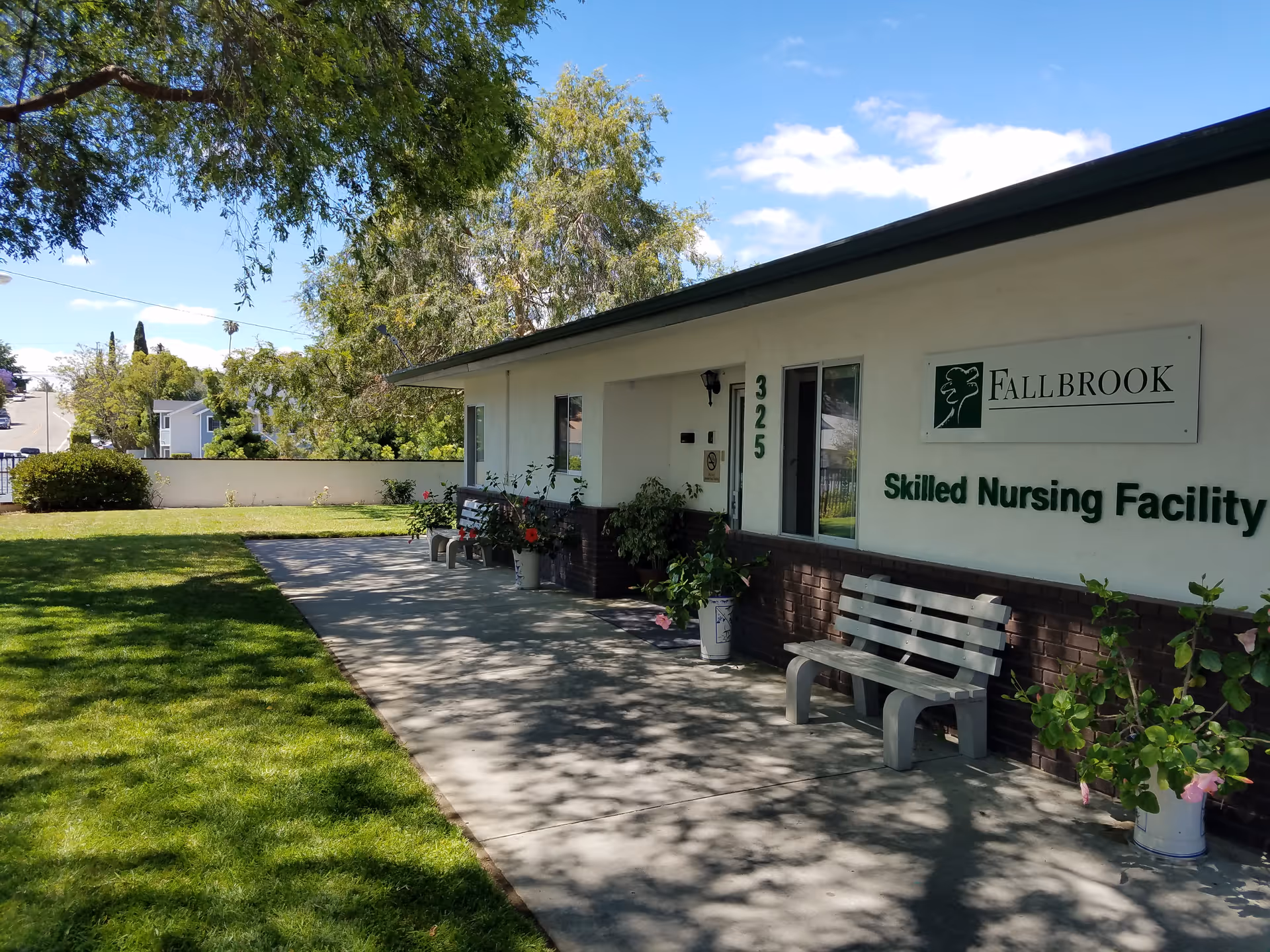 Exterior view of Fallbrook Skilled Nursing Facility building with a sidewalk, benches, potted plants, and a grassy lawn under a partly cloudy sky.
