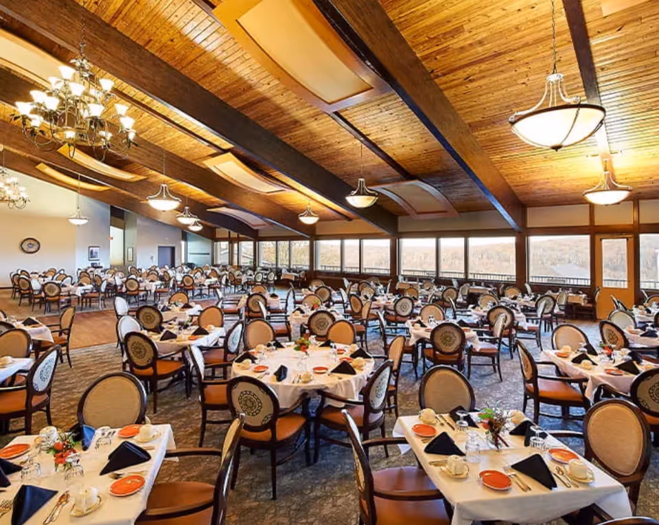 A large dining room with multiple round and rectangular tables set with white tablecloths, orange plates, black napkins, cups, and silverware. The room features a high wooden ceiling with exposed beams and several chandeliers and pendant lights. Large windows line one side of the room, offering a view of the outdoors.
