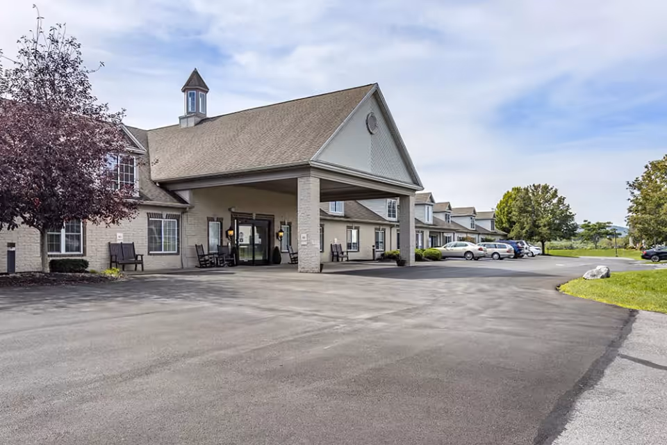 Exterior view of a senior living facility building with a large covered entrance, several parked cars, and trees around the parking lot under a partly cloudy sky.