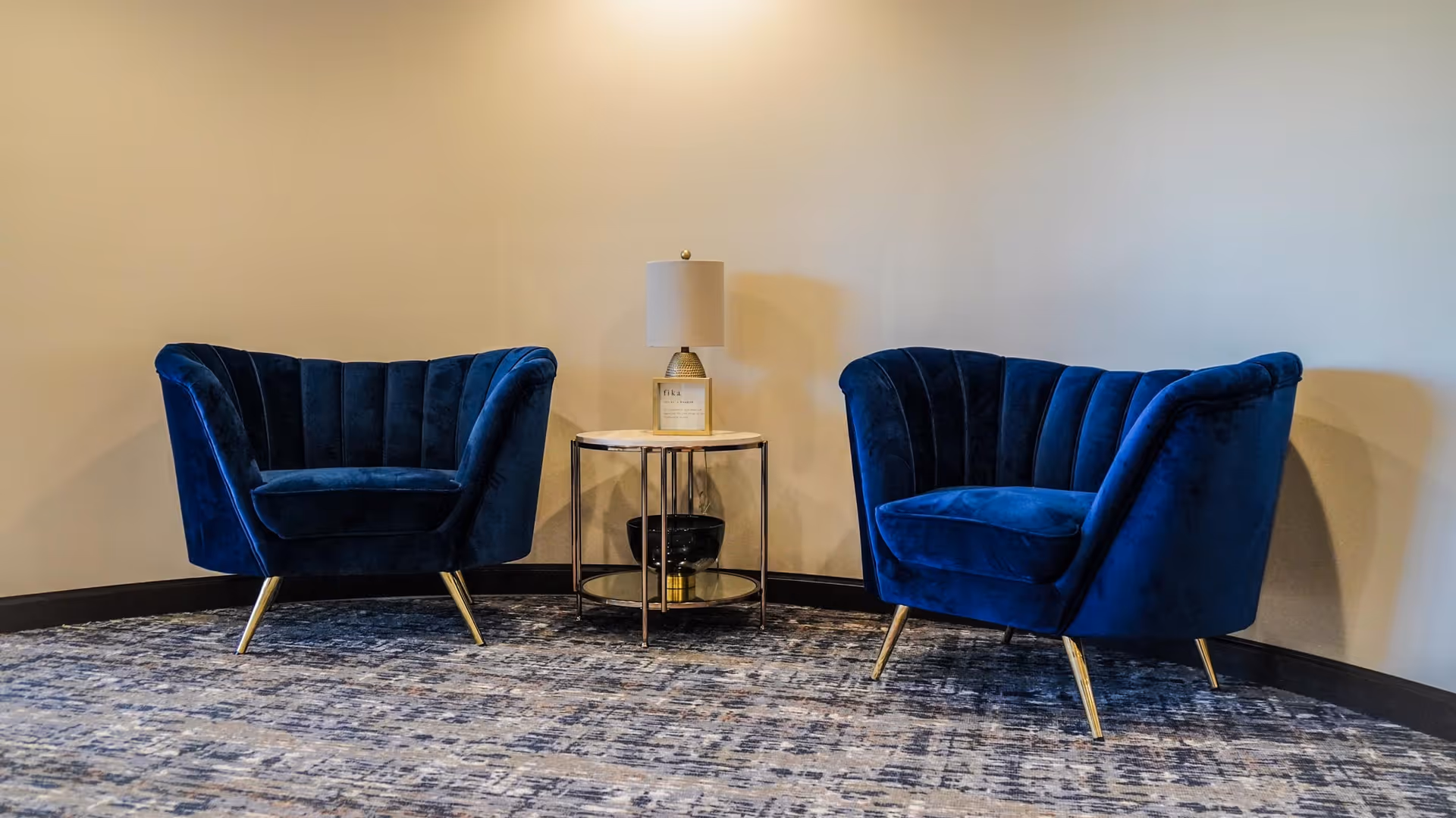 Two blue velvet armchairs with gold legs positioned on a patterned carpeted floor, separated by a small round side table with a lamp and decorative items, against a plain beige wall.