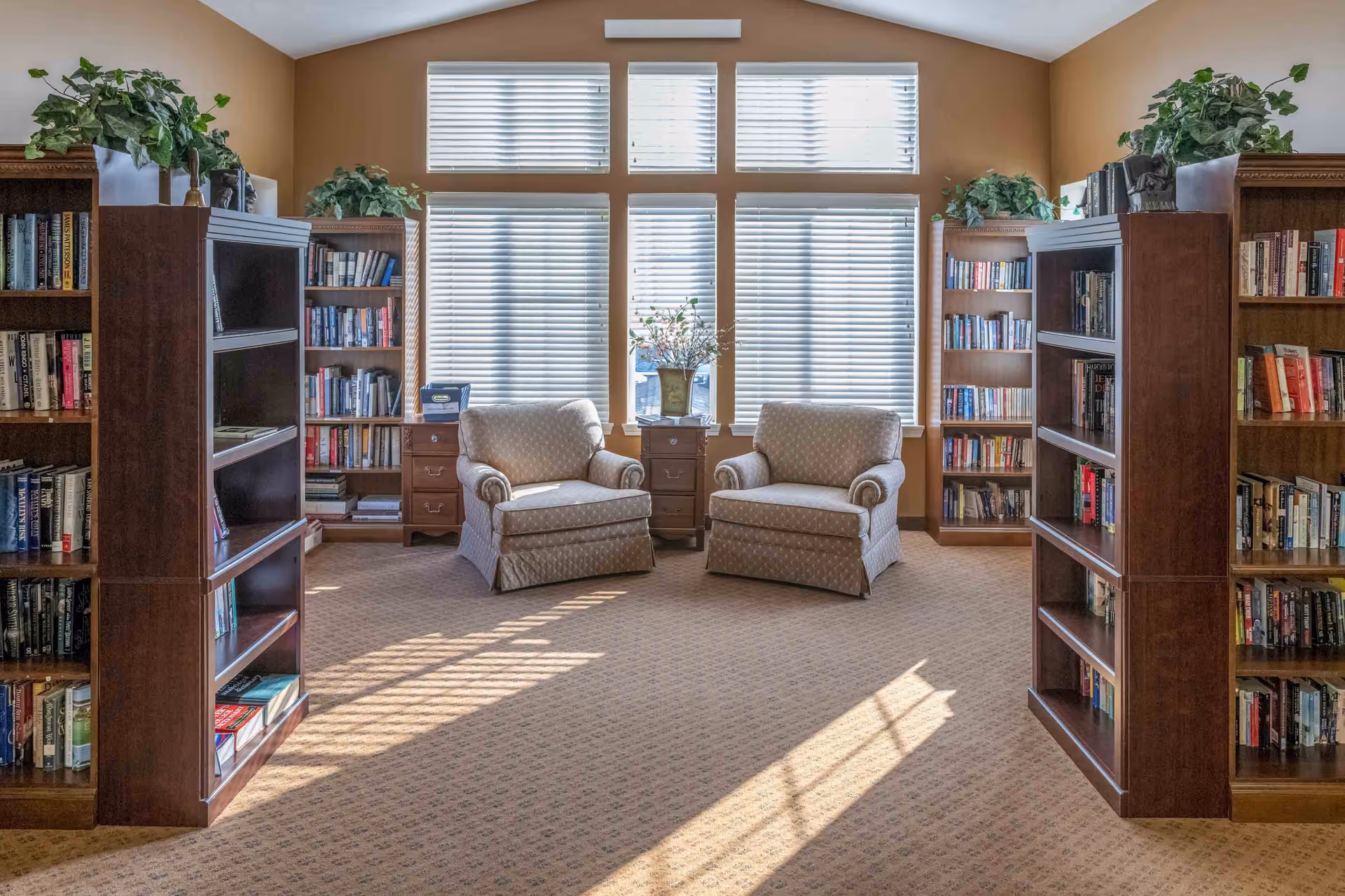 Sunlit reading room with bookshelves and two upholstered armchairs in front of large windows.